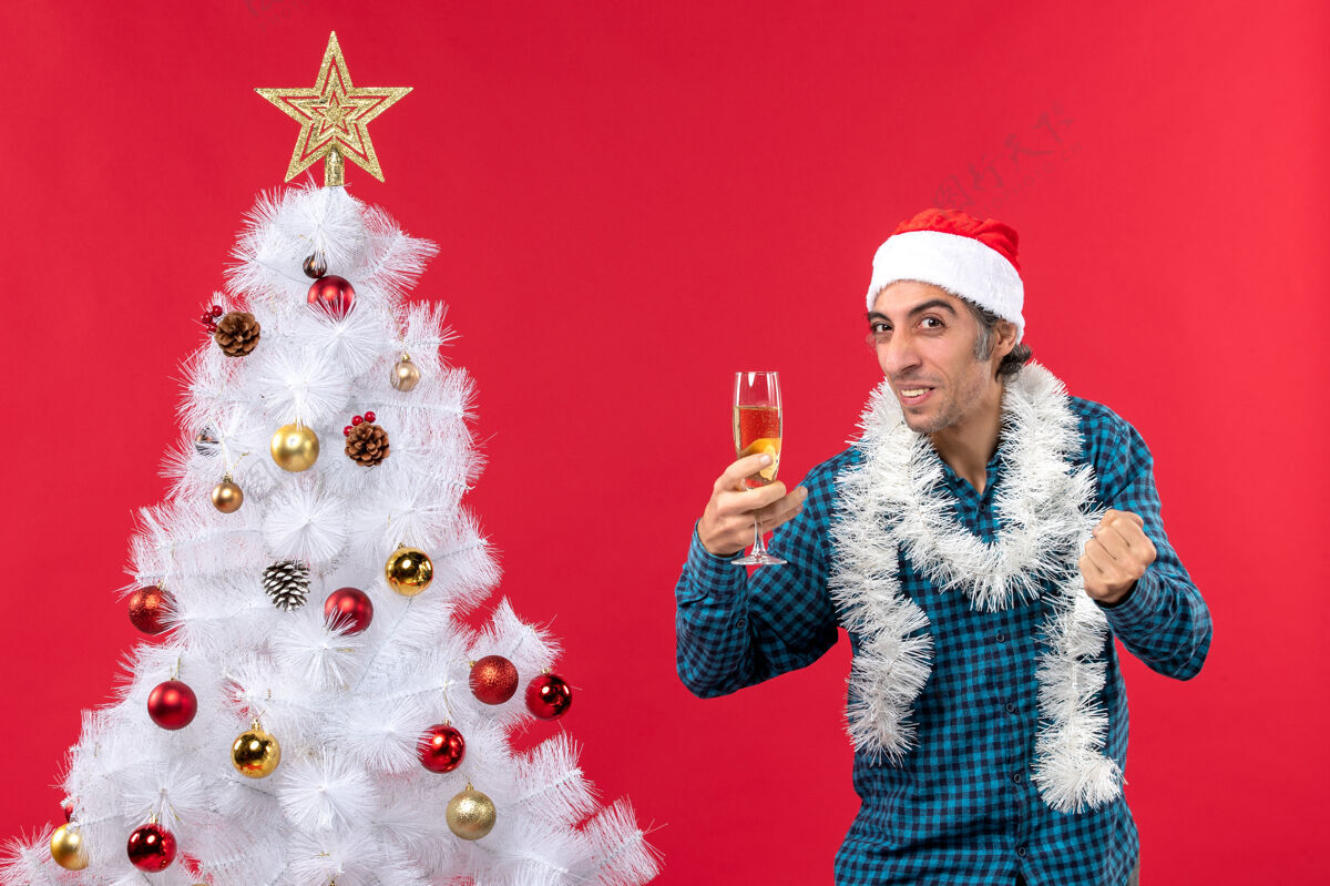 搞笑的圣诞节心情与情绪有趣的年轻人与圣诞老人帽子在一个蓝色的衬衫剥离举行一杯葡萄酒在圣诞树附近抬头看新的圣诞老人举行