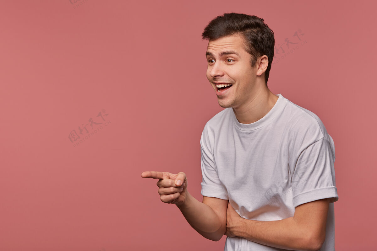 抬高一幅室内肖像画 一位快乐的年轻黑发男性 兴高采烈地站在一旁 举手用食指指着 穿着灰色t恤站在粉色背景上食指发型微笑