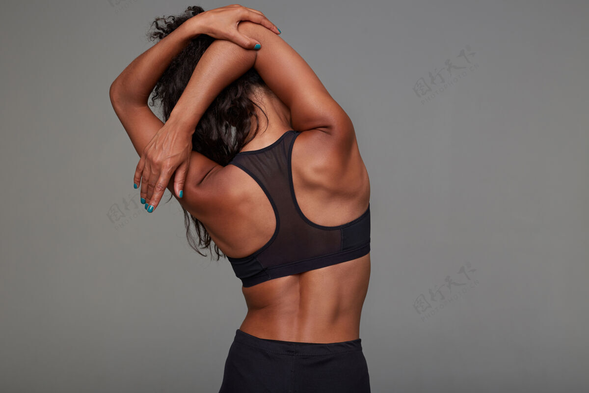 黑发年轻的运动型深色皮肤卷曲深色皮肤女性伸展双手 同时摆出黑色运动上衣的后视图健身女模特运动表情衣服