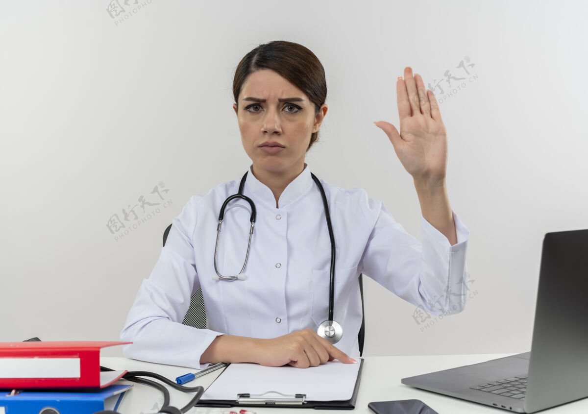 手势严格的年轻女医生 穿着医用长袍 带听诊器 坐在办公桌旁 在电脑上工作 带医疗工具 显示停止手势 留有复印空间穿着医生坐着