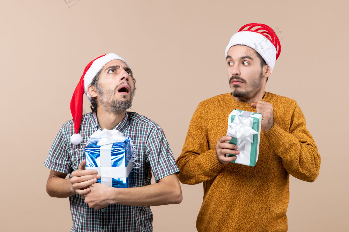 男性前视图两个困惑的家伙戴着圣诞帽 拿着圣诞礼物在米色孤立的背景圣诞老人困惑帽子