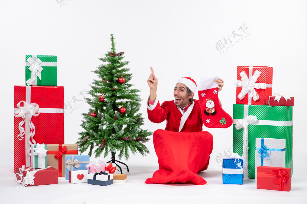 惊讶惊喜的圣诞老人正指着上面坐在地上 拿着圣诞袜靠近礼物和装饰在白色背景上的新年树圣诞老人礼物帽子