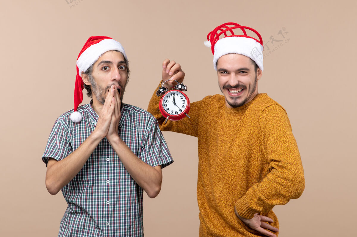 圣诞节前视图两个圣诞节男人一个惊喜和一个快乐的人拿着一个闹钟在米色孤立的背景帽子米色男人