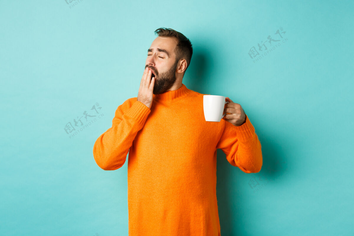 胡须昏昏欲睡的帅哥喝着咖啡打着哈欠 穿着橘色毛衣站在浅蓝绿色的墙上胡须英俊饮料