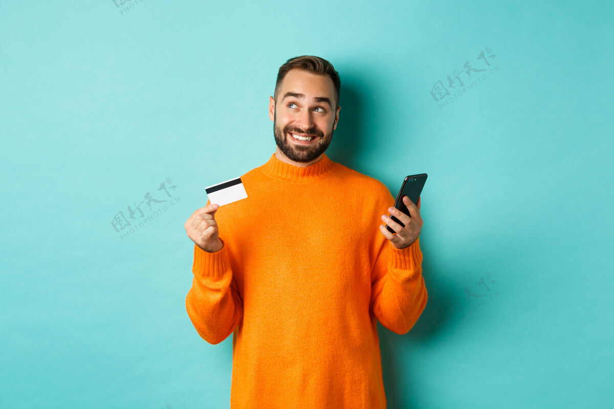 信用网上购物帅哥思考 拿着信用卡的智能手机 在网店付款 站在浅绿的墙上黑发应用程序时髦