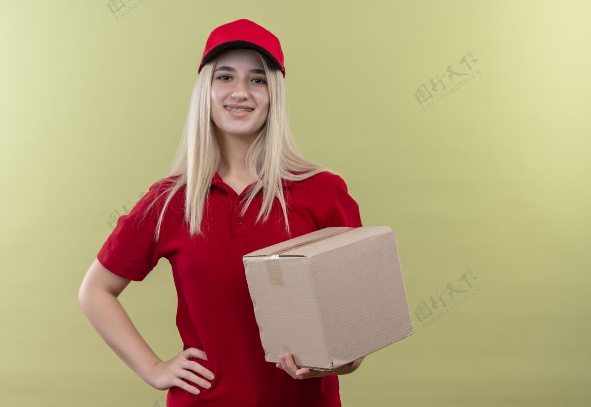 抱着带着微笑的小女孩穿着红色t恤 戴着帽子 戴着牙套 在绿色的背景下拿着盒子年轻红色盒子