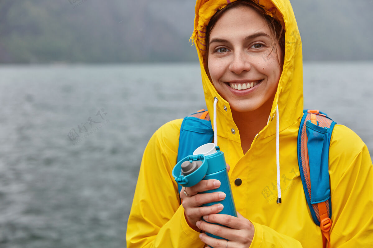 烧瓶欢喜女子的后景穿着带兜帽的黄色雨衣 开心地笑着 走在山湖的岸边单独背包热