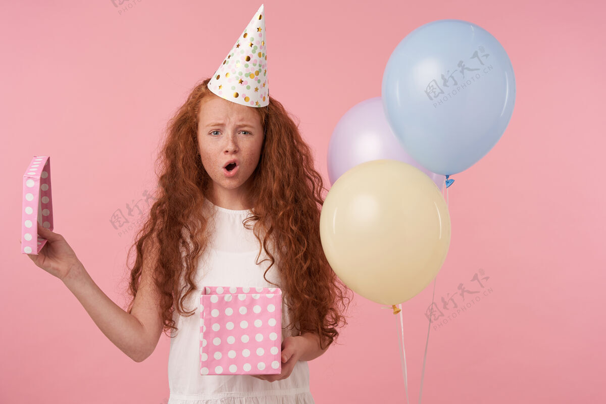 举行室内拍摄的红发女孩 卷发 白色连衣裙 戴着生日帽 站在粉色摄影棚背景下 愁眉不展地看着镜头 手里拿着礼盒气球情绪化打开包装