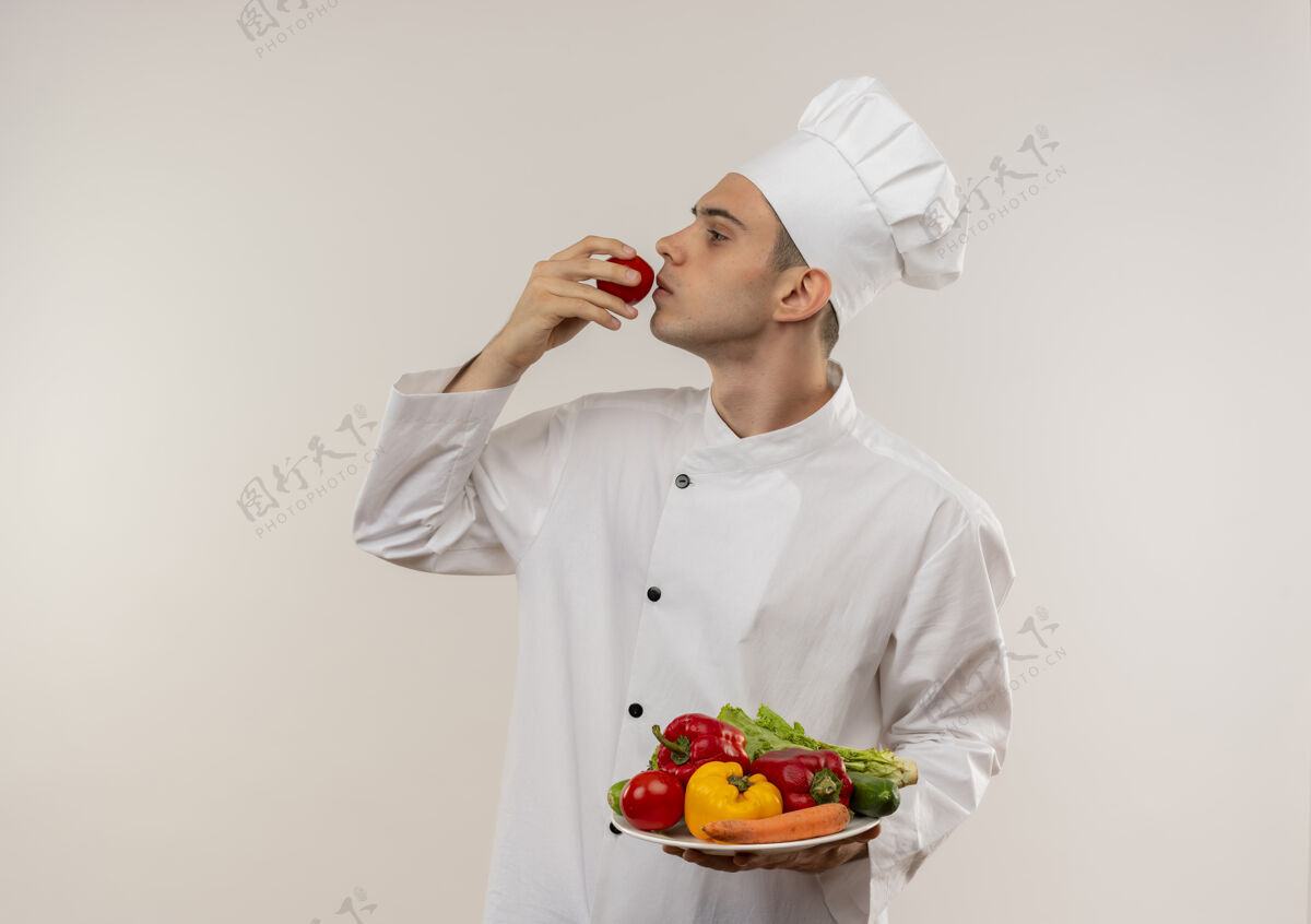 厨师站在侧视图年轻的男厨师穿着厨师制服拿着盘子上的蔬菜和闻西红柿在他的手与复制空间轮廓嗅着制服