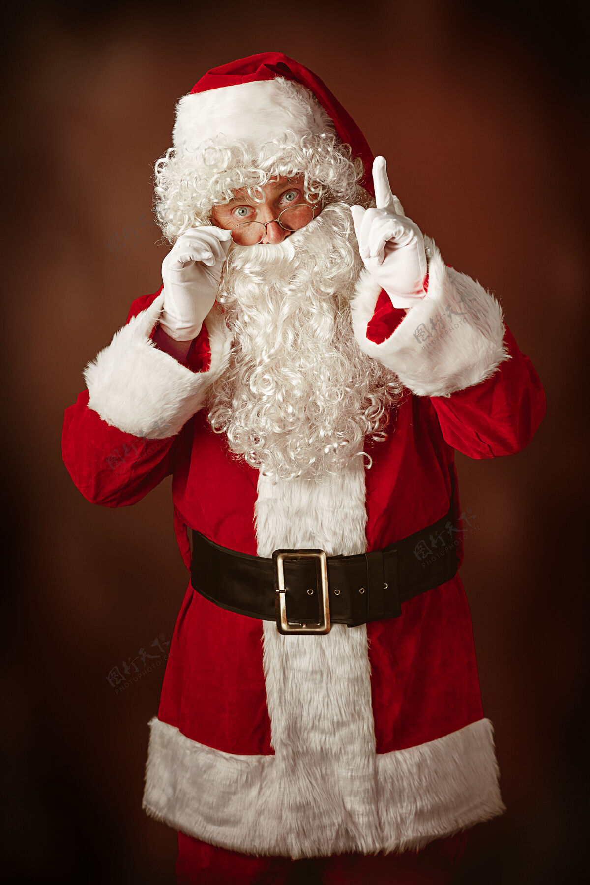 男性穿着圣诞老人服装的人的肖像-一个豪华的白胡子 圣诞老人的帽子和红色的服装在红色工作室的背景手老人