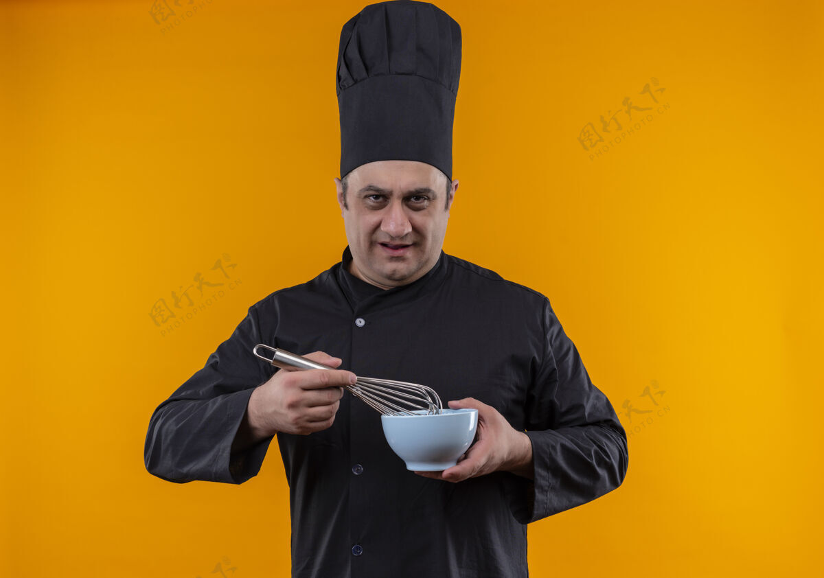 厨师严格的中年男性厨师 身着厨师制服 手持黄色背景的搅拌器和碗严格碗拂