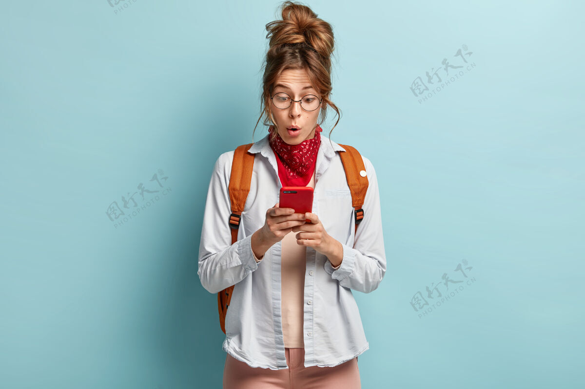 电话震惊的年轻潮人女孩惊讶地看着手机 收到意想不到的信息 背着背包持有使用眼镜