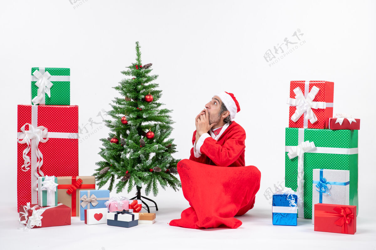 装饰吓了一跳的年轻人打扮成圣诞老人 拿着礼物和装饰好的圣诞树坐在白色背景的地上年轻人圣诞礼物