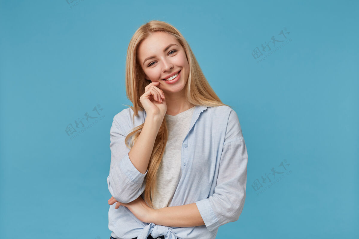 女士积极的金发长发女士穿着蓝色衬衫和灰色t恤休闲服摆姿势 微笑迷人的样子和感动的脸举起手来女人手抚摸