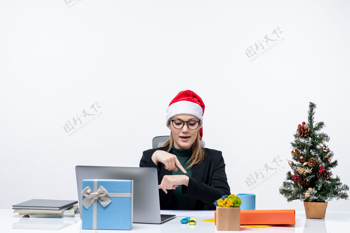迷人的女人一位年轻漂亮的女士戴着圣诞老人帽坐在桌子旁 桌上放着圣诞树和礼物 她正在检查自己在办公室的时间圣诞老人坐着圣诞老人