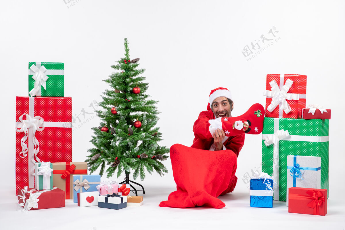 地有趣的圣诞老人坐在地上 穿着圣诞袜 靠近礼物和装饰在白色背景上的新年树圣诞老人礼物季节性