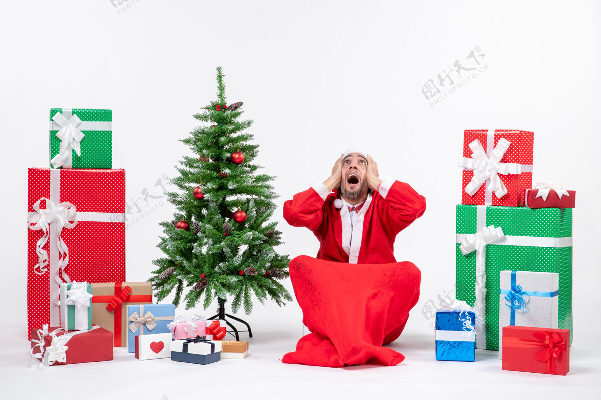 年轻人紧张的年轻人打扮成圣诞老人和礼物装饰圣诞树坐在地上看上面的白色背景圣诞树圣诞老人装饰