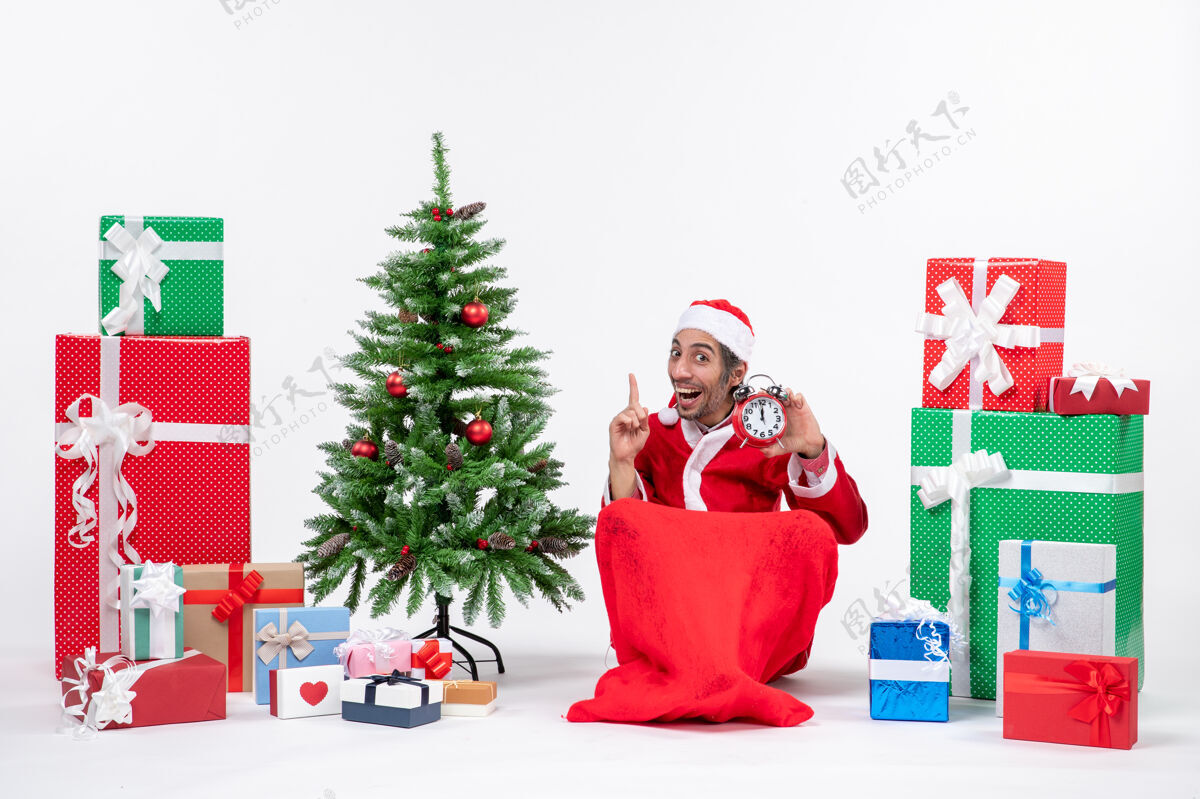 微笑微笑有趣的年轻人庆祝新年或圣诞节假期坐在地上 拿着时钟指向上方的礼物和装饰圣诞树礼物地年轻人
