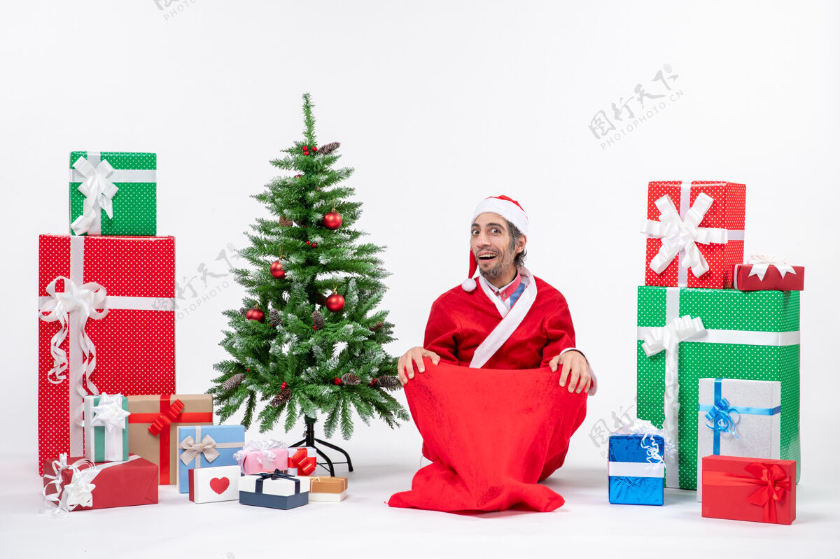 装饰震惊的年轻人打扮成圣诞老人与礼物和装饰圣诞树坐在地上的白色背景圣诞老人礼物地
