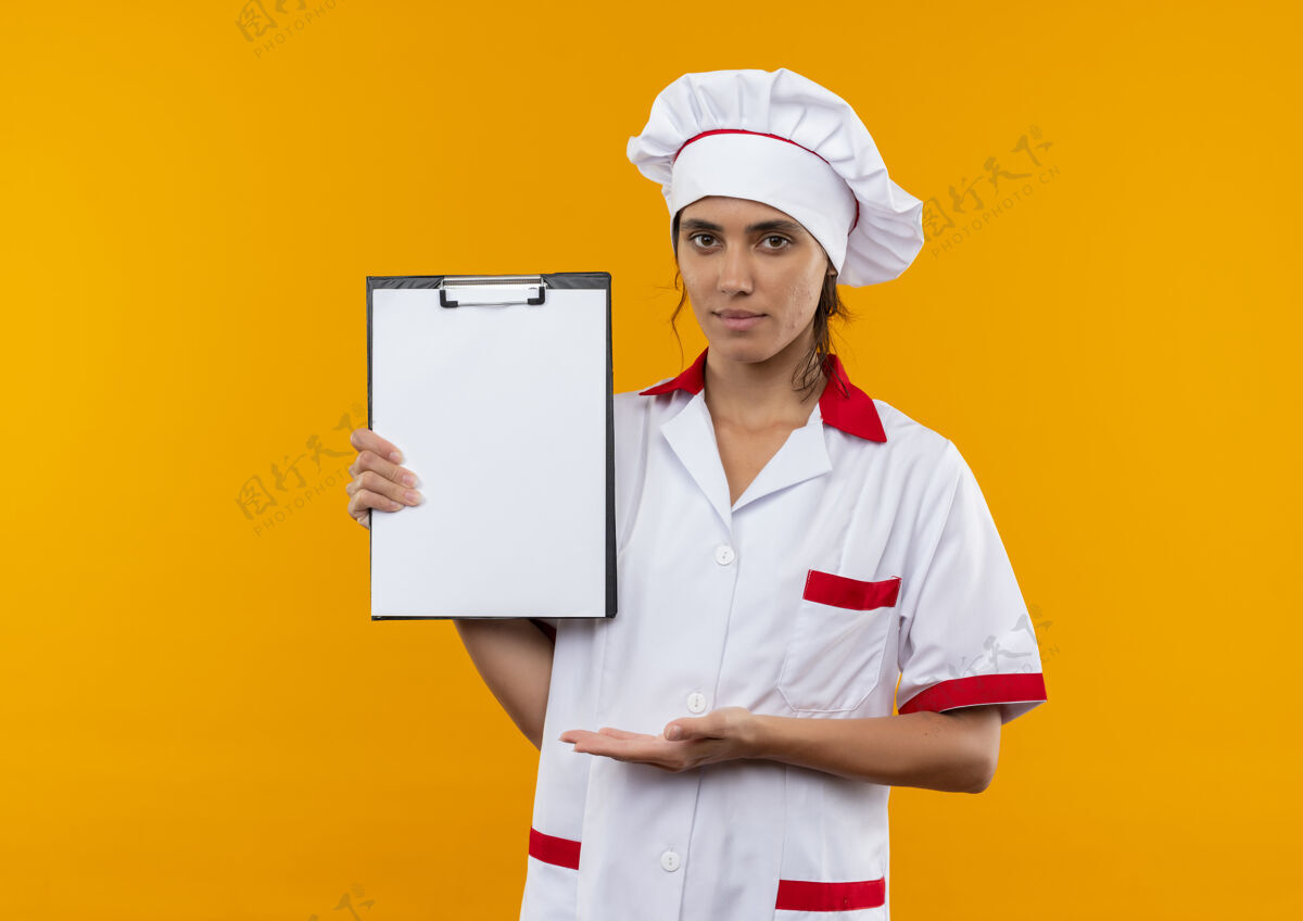 制服高兴的年轻女厨师穿着厨师制服举行 并显示与手剪贴板上孤立的黄色墙壁与复制空间显示厨师年轻