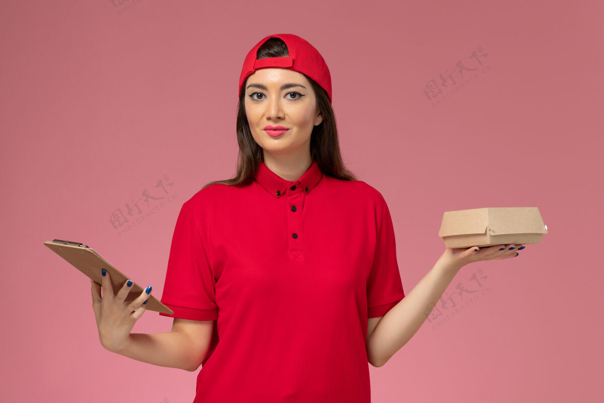 制服正面图：身穿红色制服斗篷的年轻女快递员 手上拿着小快递食品包和记事本放在粉红色的墙上记事本年轻女快递肖像
