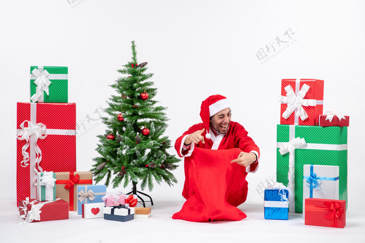 男人笑容可掬的年轻人打扮成圣诞老人 拿着礼物和装饰好的圣诞树坐在白色背景的地上年轻人帽子装饰