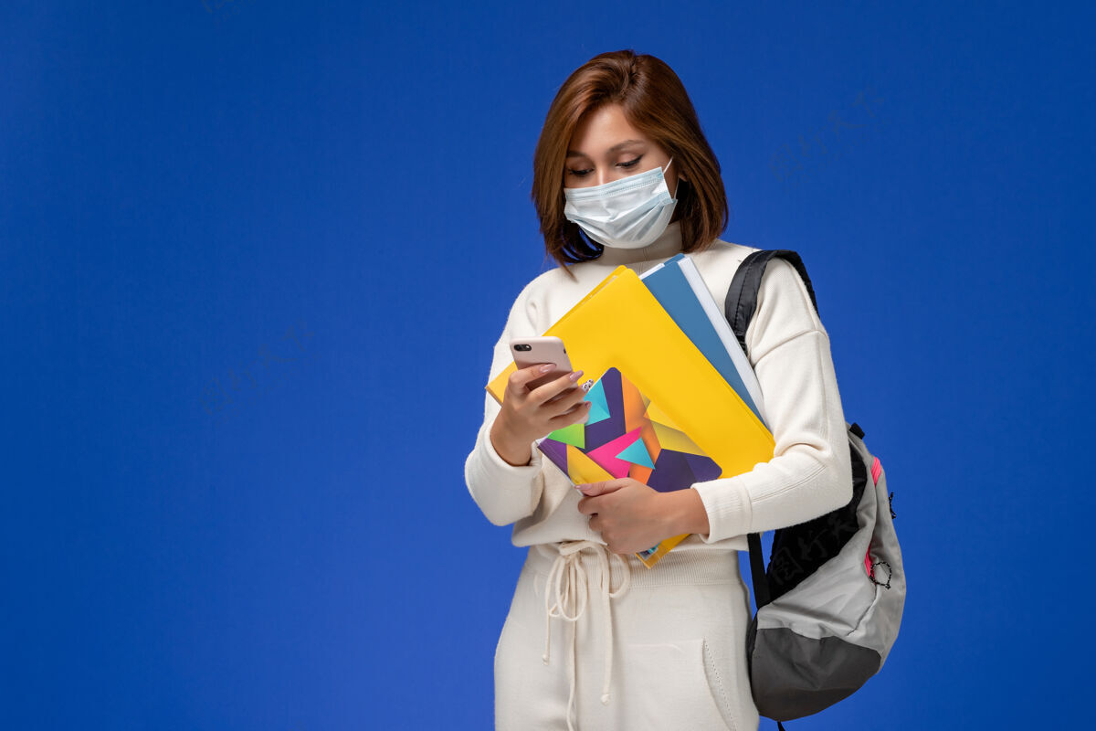 面具正面图穿着白色球衣的年轻女学生戴着带包的面具和抄写本 在蓝色的墙上用手机文案学生使用