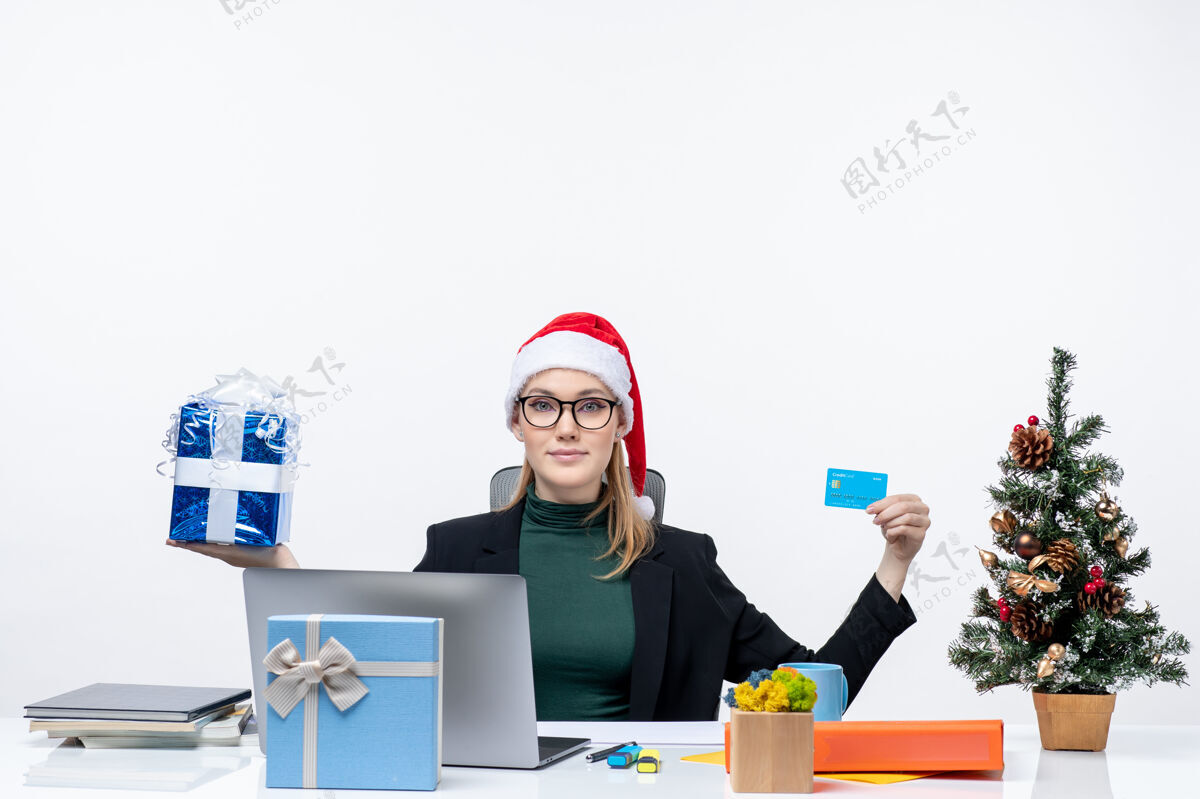 圣诞节戴着圣诞老人帽戴着眼镜的快乐年轻女子坐在一张桌子旁 白色背景上展示圣诞礼物和银行卡电脑专业年轻女人