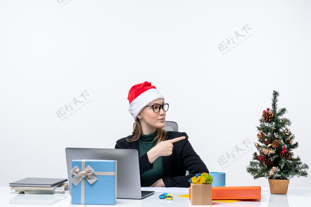 什么新年的气氛与金发女人坐在一张桌子上圣诞树和圣诞老人帽子上的礼物指向在白色背景的左边的东西金发帽子女人