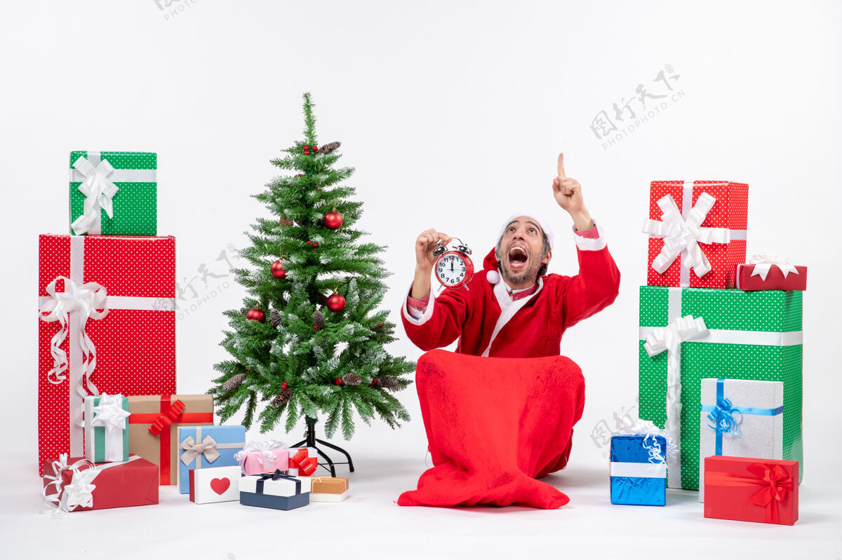 礼物紧张兴奋的圣诞老人指着上面坐在地上 在礼物和装饰圣诞树旁的白色背景上展示时钟坐着人礼物