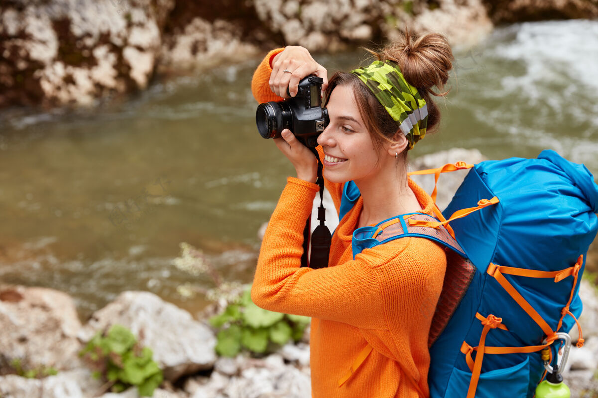 爱好欢笑的女游客用相机拍照 聚焦到远处 戴着头巾 穿着橙色毛衣 欣赏大自然的美景小溪自然摄影