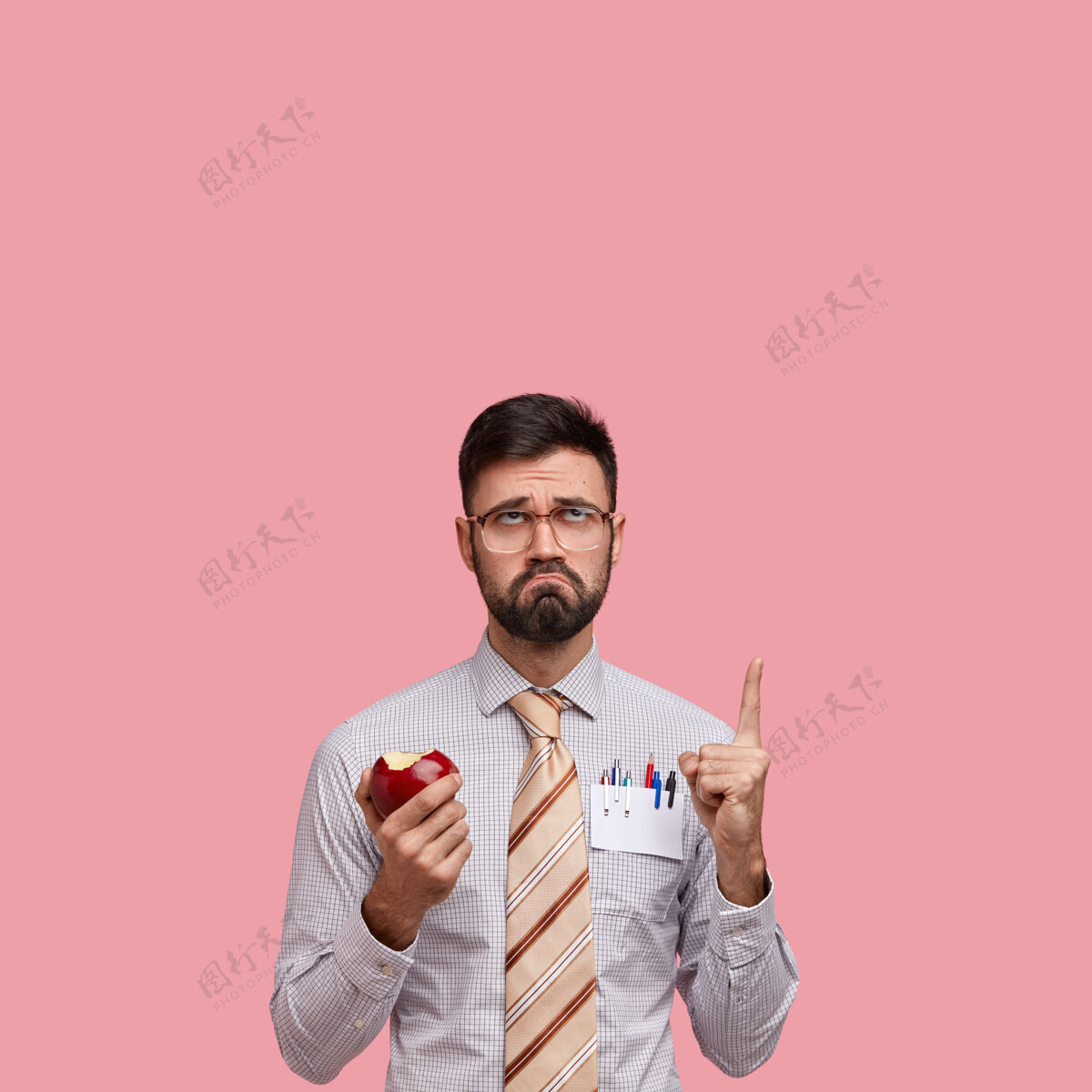 皱眉不开心男人的垂直镜头 留着深色的胡茬 表情消极 吃着多汁的苹果 穿着正式的衣服 食指朝上指着个性企业家聪明