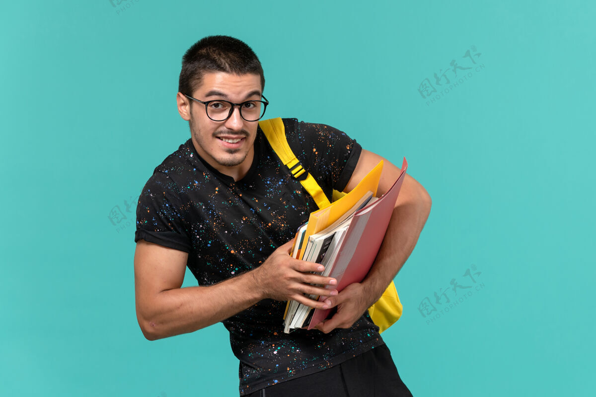 模特身穿深色t恤黄色背包的男学生正面图 浅蓝色墙上有文件和书籍正面微笑人