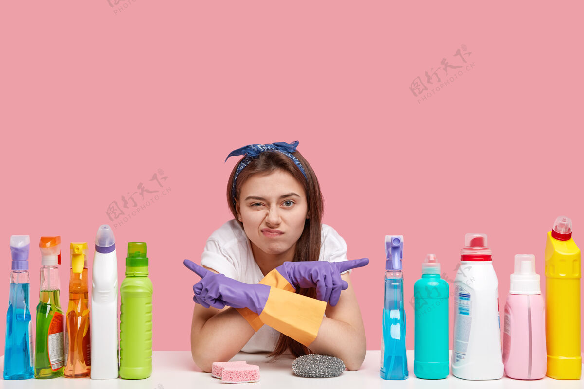 墙不高兴的女人指向不同的侧面 展示清洁用品 不喜欢效果 戴着紫色的橡胶手套清洁剂女佣负面