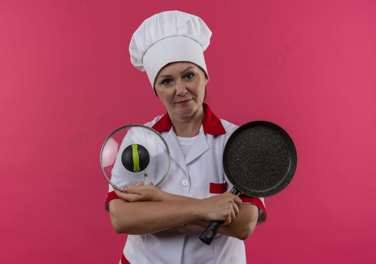 看着身着厨师制服的中年女厨师手拿煎锅和锅盖 放在隔离的粉色墙壁上 有复印空间中年人平底锅煎炸