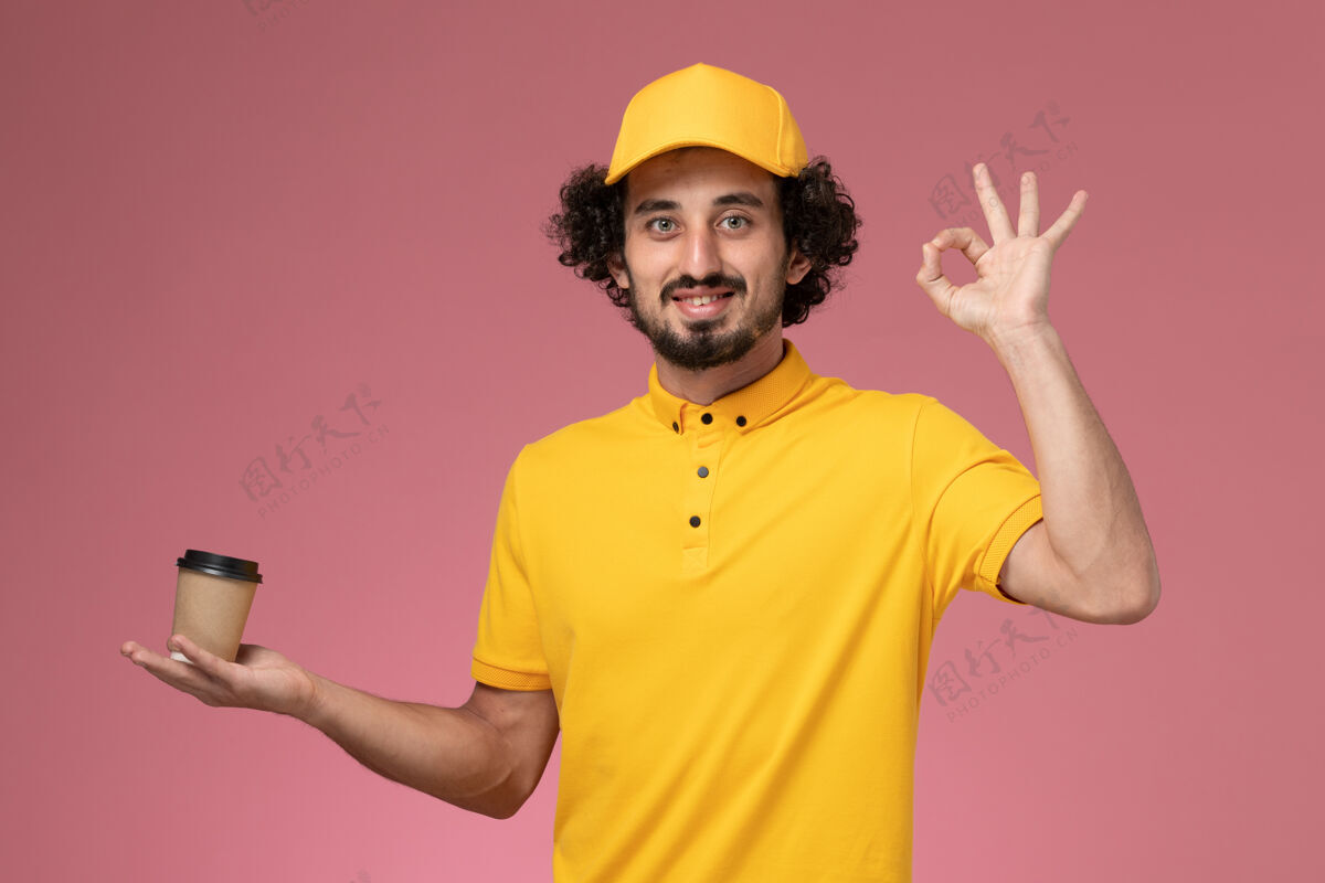 工作正面图：身穿黄色制服 披风的男性快递员在粉色墙上拿着送货咖啡杯服务人帽子