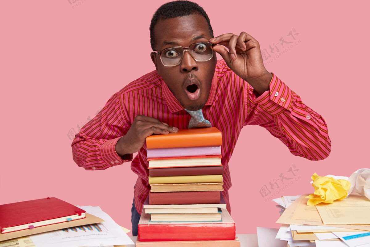 男性一个令人惊讶的黑人年轻人透过眼镜仔细地看了看 倚在一堆书上 手放在眼镜边上文学桌面情绪化