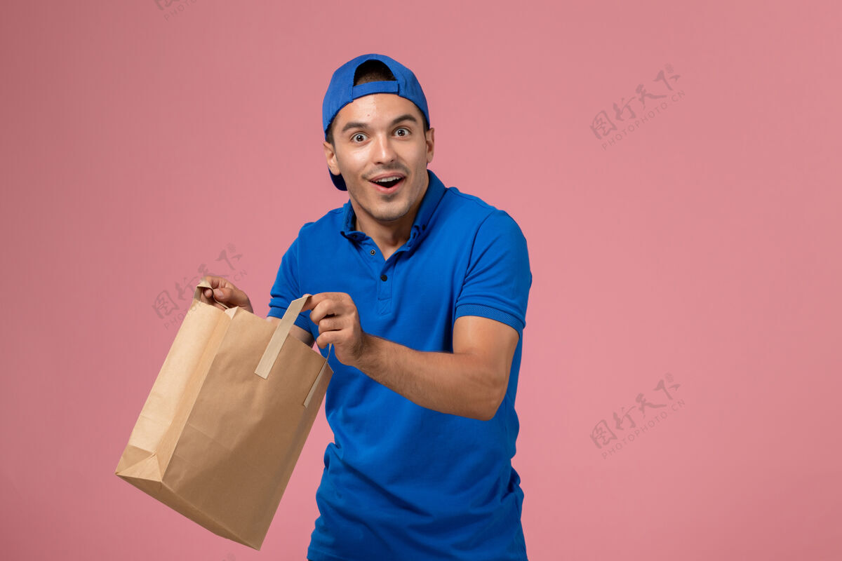 男孩正面图：身穿蓝色制服和斗篷的年轻男性快递员 手上拿着快递纸包裹 放在粉红色的墙上服务手微笑