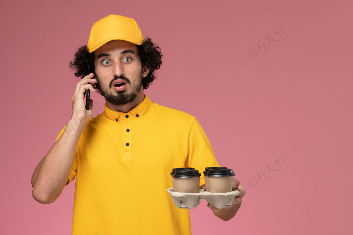 服务正面图穿着黄色制服的男信使拿着棕色的咖啡杯在粉红色的墙上打电话男性工作工作