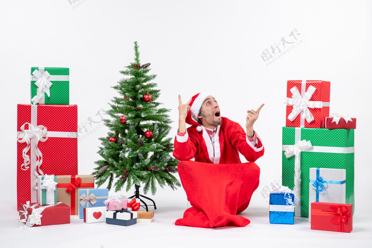 看困惑的年轻人打扮成圣诞老人与礼物和装饰圣诞树坐在地上看上面的白色背景年轻人装饰年轻人
