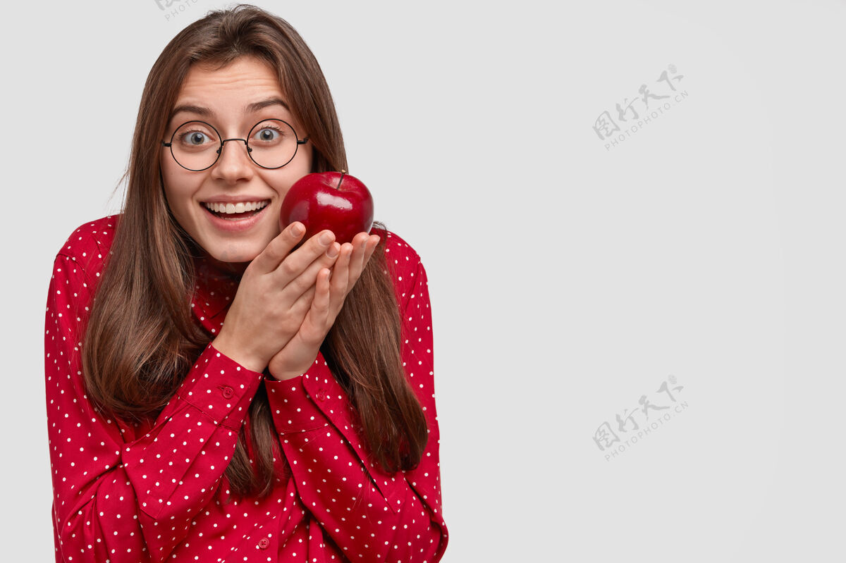 高兴笑容满面的欧洲女人 捧着红苹果 穿着时髦的衣服 戴着圆眼镜 喜欢吃水果黑发眼镜水果