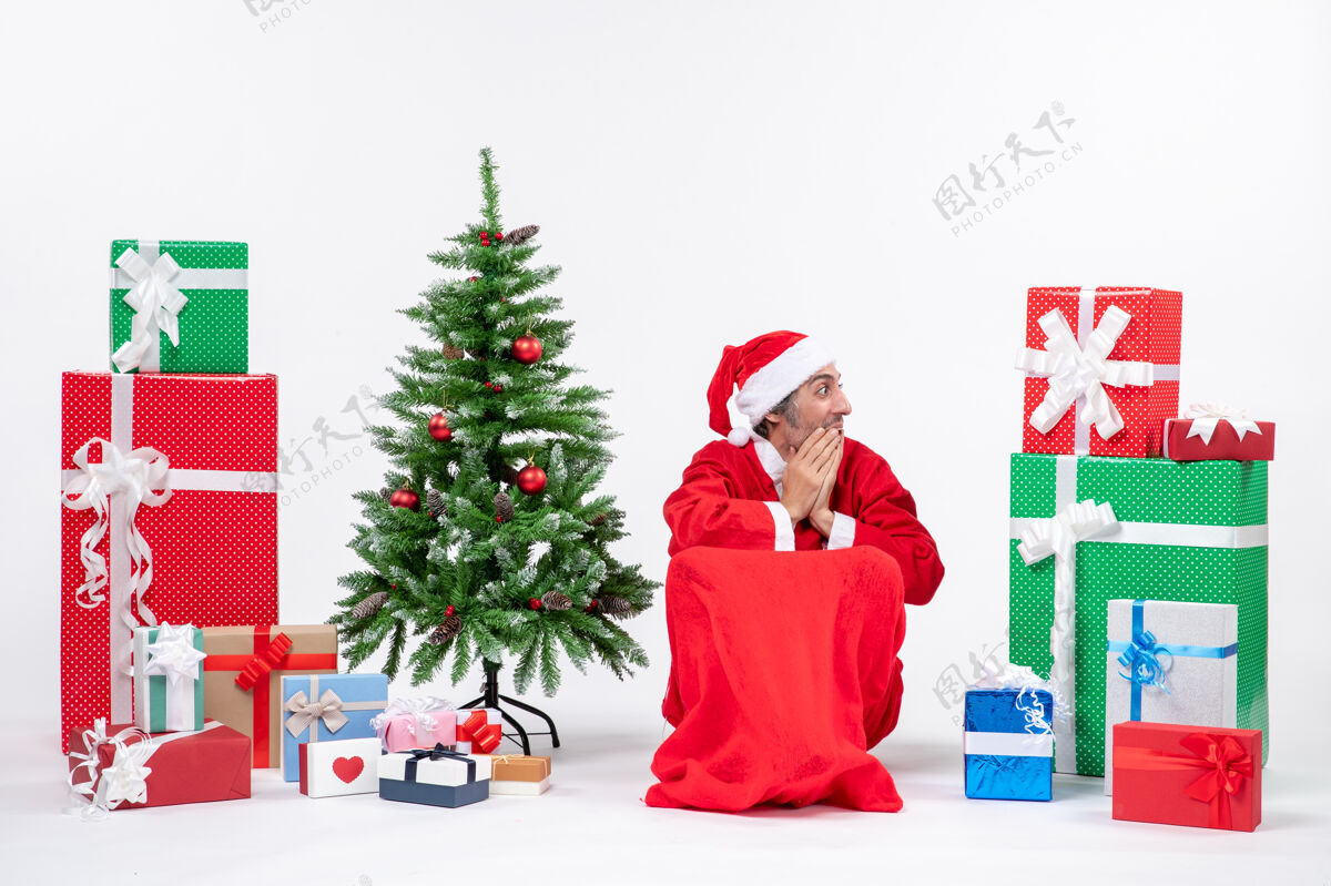 圣诞树情绪激动的年轻人打扮成圣诞老人 拿着礼物和装饰过的圣诞树 看着左边某个地方的白色背景看兴奋情绪