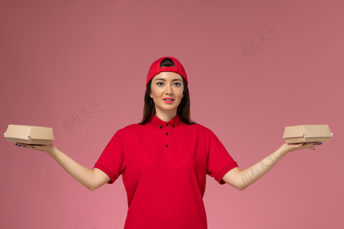 人正面图：身穿红色制服和斗篷的年轻女快递员 手上拿着小包裹 放在粉红色的墙上女人年轻漂亮