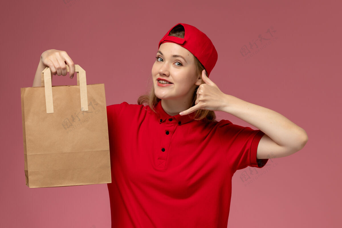 视图正面图：身穿红色制服和披风的年轻女快递员手持快递食品包 微笑着站在粉红色的墙上女性肖像年轻女快递