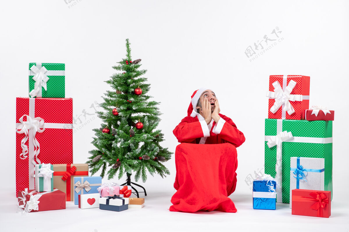打扮情绪激动的关心的年轻人打扮成圣诞老人与礼物和装饰圣诞树上的白色背景镜头圣诞老人圣诞树圣诞老人