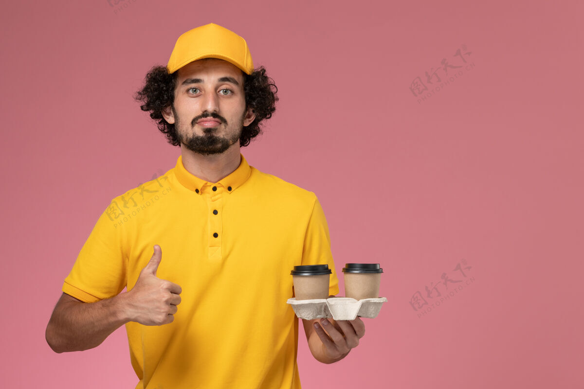咖啡正面图：身穿黄色制服 披风的男性快递员在粉色墙上拿着送货咖啡杯人正面男士