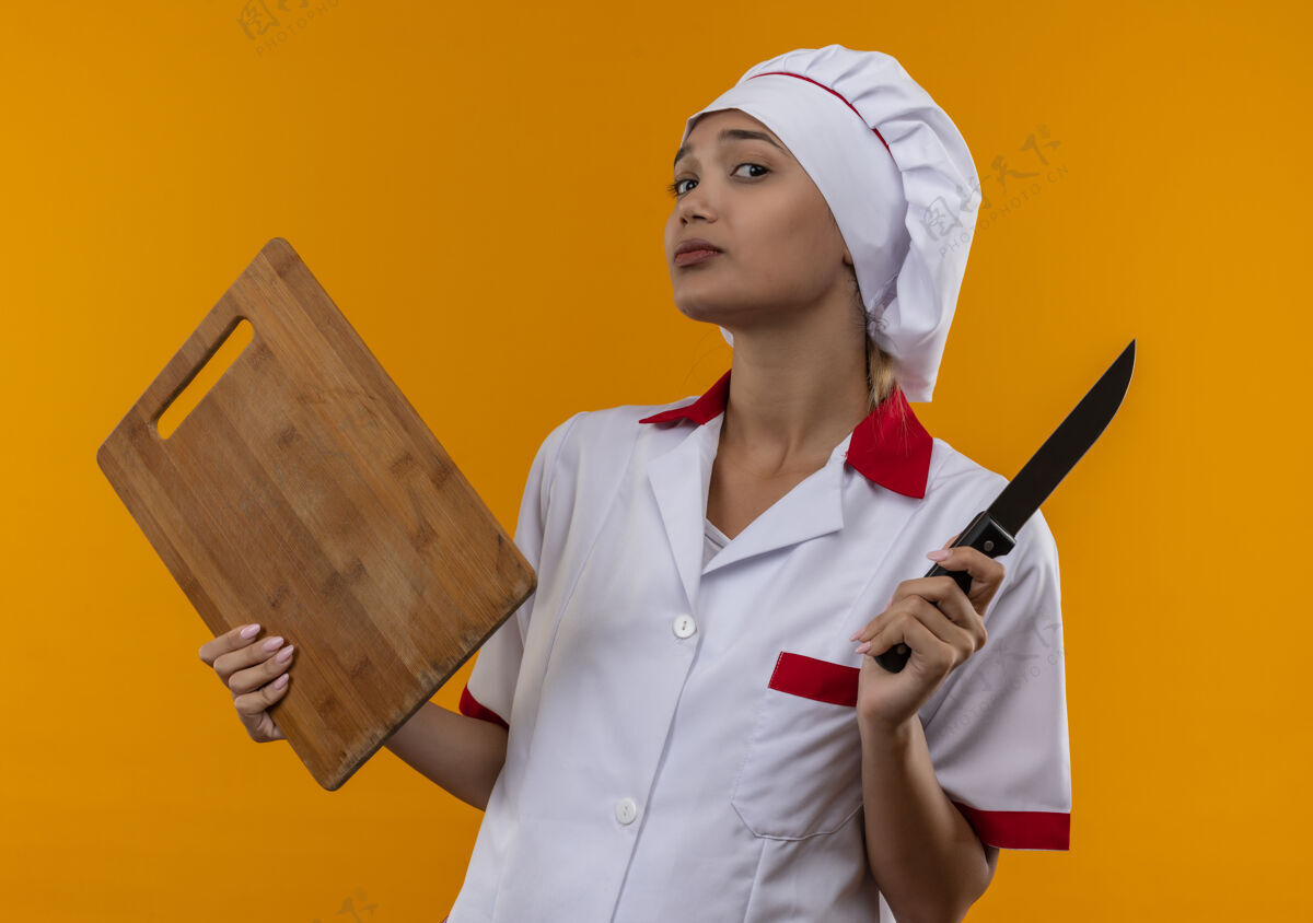 厨师年轻的女厨师穿着厨师制服拿着砧板和刀站在隔离的橙色墙上穿年轻制服