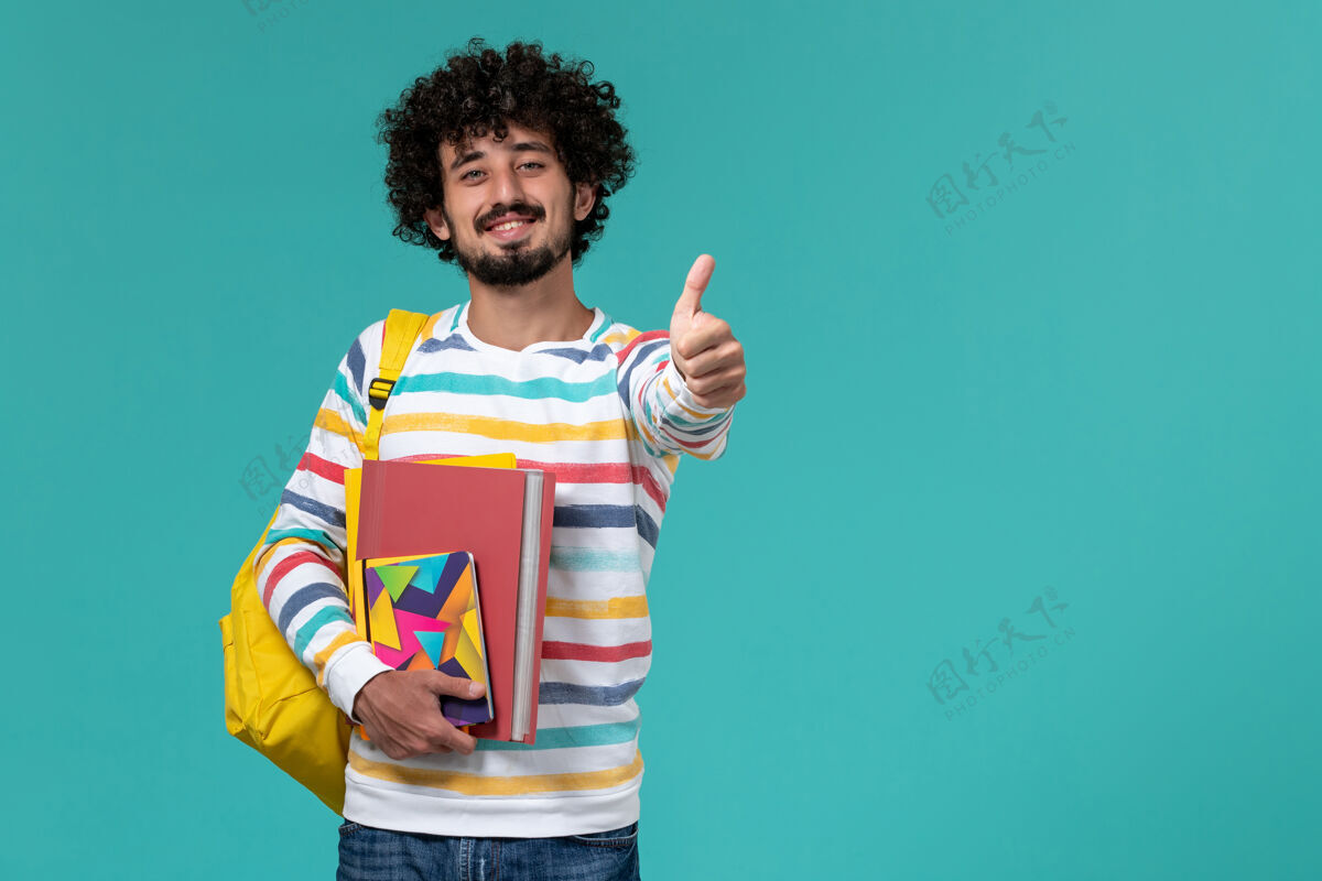 衬衫身着彩色条纹衬衫 背着黄色背包 拿着文件和复印件的男学生在蓝色墙上微笑的正面视图男学生彩色微笑